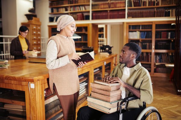 Portrait of young African-American man in wheelchair holding stack of books and chatting with friend at college library
