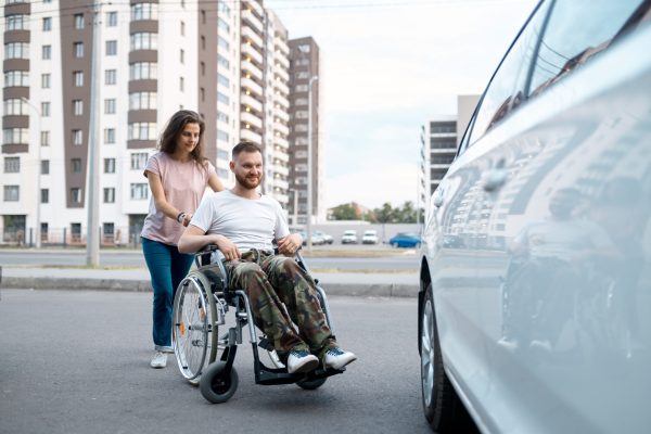 Young family couple with wheelchair walking to a car. Paralyzed veteran and disability, care for a disabled man. Husband and wife overcome difficulties together